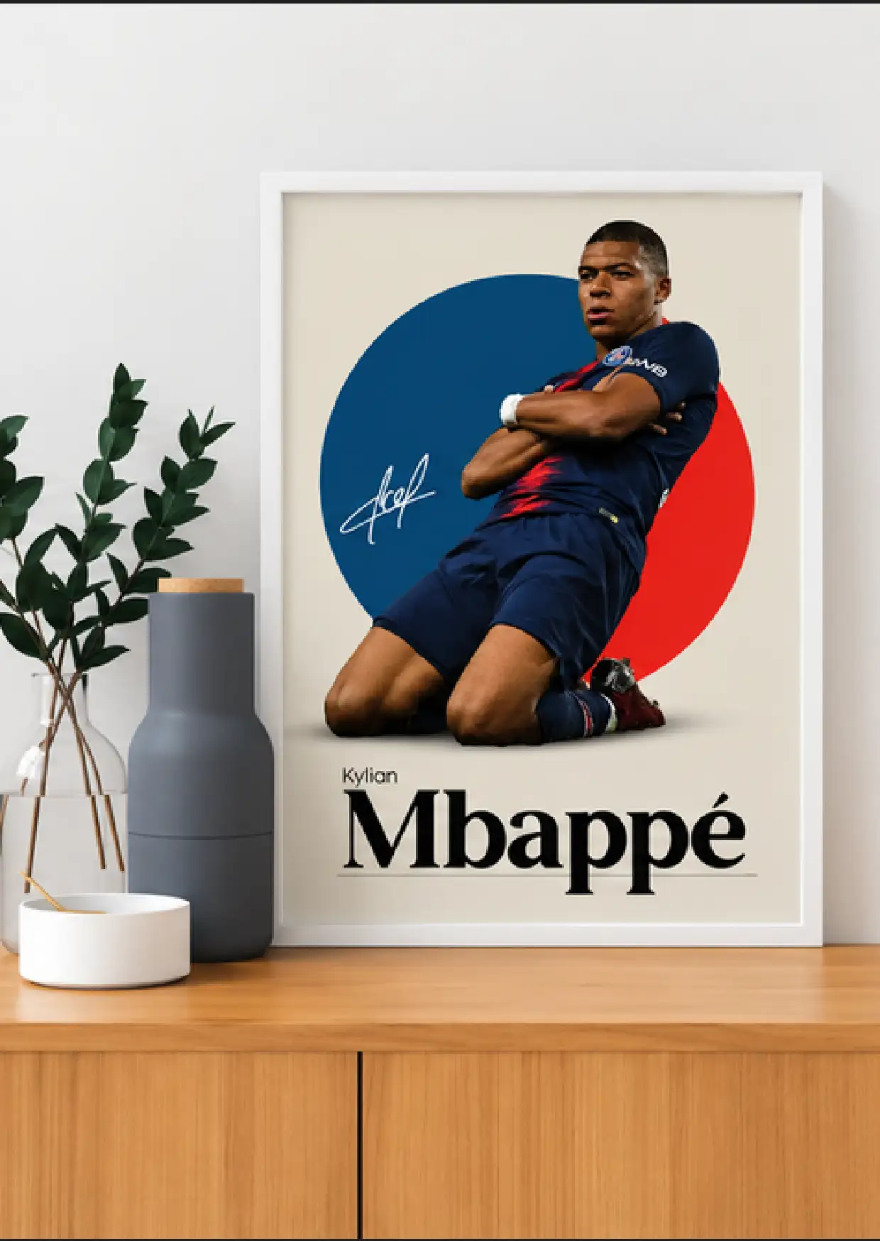 White-framed Kylian Mbappé poster showing a crossed-arm knee-slide celebration against a blue and red circle on cream.